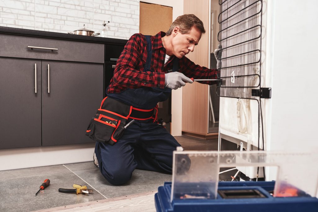 A repairman fixing a fridge from the back, using a screwdriver